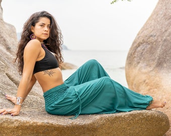May include: A woman wearing a black crop top and teal pants sits on a large rock. She has long dark hair and is looking to the right. The rock is in front of a body of water.