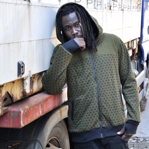 May include: A man wearing a green and black patterned hooded jacket with a zipper. He is standing next to a large lorry.