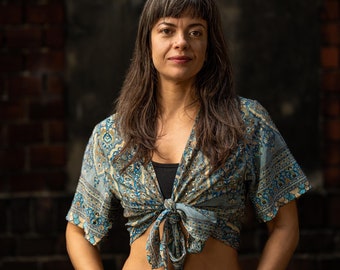 May include: A woman with long brown hair is wearing a blue and white patterned top with a knotted front. The top is tied at the waist, revealing her midriff. She is standing in front of a brick wall.