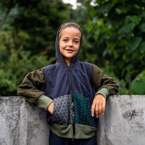 May include: A young person wearing a blue, green and grey hooded jacket with a zip. The jacket has a geometric pattern on the front. The person is standing in front of a concrete wall.