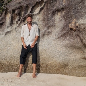 May include: A man wearing a white linen shirt with a long pendant necklace and black trousers stands in front of a large rock formation.
