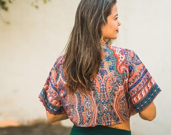 May include: A woman with long brown hair is wearing a colorful paisley print top with a green skirt. She is standing in front of a white wall.