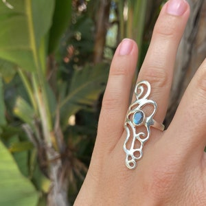 Könnte beinhalten: Ein silberner Ring mit einem filigranen Design und einem zentralen, oval geformten, blau-grünen Edelstein. Die dekorativen Wirbel und Kurven des Rings erzeugen einen eleganten, künstlerischen Look. Der Ring wird am Finger getragen.