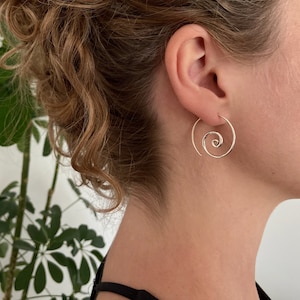 Könnte beinhalten: Silberne Spiral-Creolen. Die Ohrringe bestehen aus einem dünnen Draht, der zu einer Spirale gedreht ist. Die Ohrringe sind an einem Ohr einer Person zu sehen.