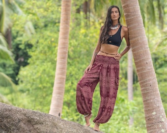 May include: A woman wearing a black crop top and red printed pants stands next to a palm tree. She is wearing a beaded necklace and a bracelet.