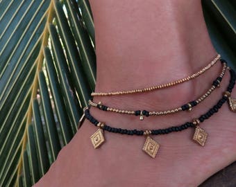 May include: Three layered anklets with gold and black beads. The top layer is a thin gold chain, the middle layer is a thicker gold chain with black beads, and the bottom layer is a black beaded chain with gold diamond-shaped charms.