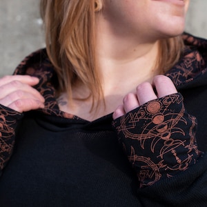 Op de afbeelding: Zwarte hoodie met lange mouwen en een bruine, hemelse print op de manchetten. De print heeft een herhalend patroon van manen, sterren en geometrische vormen.