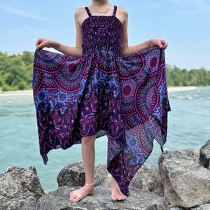 以下が含まれることがあります： 紫と青の模様のドレス。シャーリング加工された胴体と流れるようなスカートが特徴です。マンダラのようなデザインで、水辺で着用されています。ドレスは非対称の裾です。