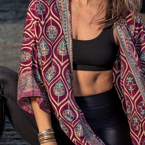 May include: A woman wearing a long-sleeved, maroon kimono with a gold and blue patterned design. The kimono is open in the front, revealing a black sports bra and black leggings.