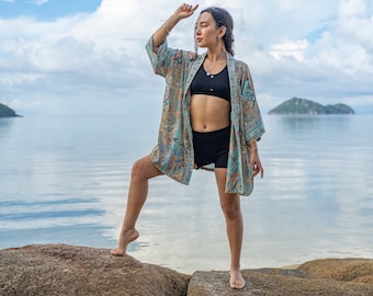May include: A woman wearing a blue and green patterned kimono with black shorts and a black sports bra stands on a rock by the ocean. The sky is blue with white clouds.