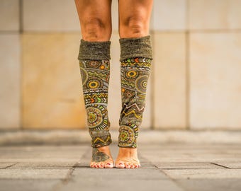 May include: A pair of olive green leg warmers with a detailed pattern of yellow, orange, green, and brown geometric shapes. The leg warmers are worn on the legs and feet of a person.