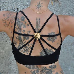 Puede incluir: Sujetador deportivo negro con un diseño de espalda único con tirantes. El sujetador presenta un cierre decorativo dorado en el centro. La modelo tiene múltiples tatuajes en la espalda y los brazos. El fondo es una pared de hormigón gris.