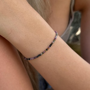 May include: A delicate bracelet made of small, colourful beads. The beads are a mix of purple, black, green, and clear, strung together to form a flexible band. The bracelet is worn on an arm.