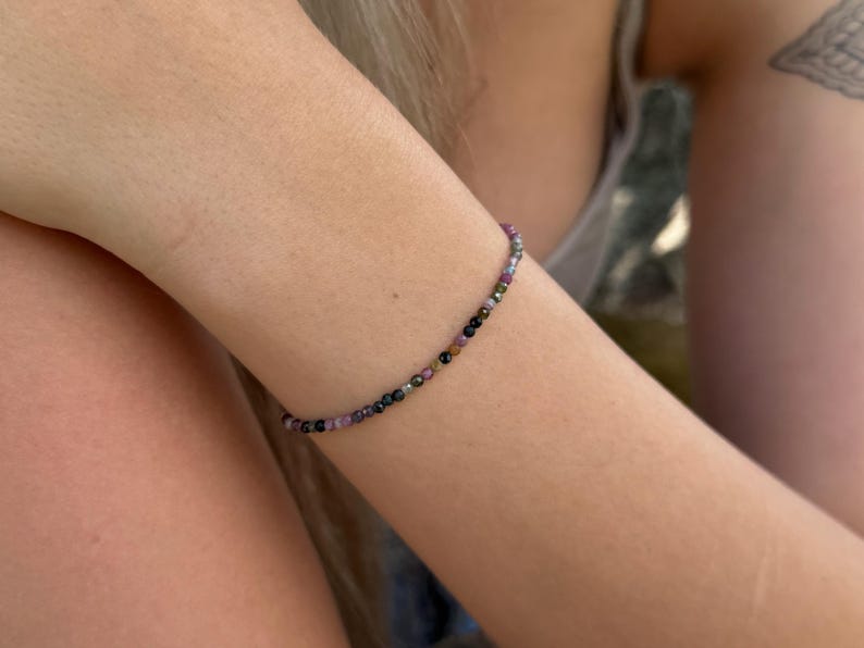 May include: A delicate bracelet made of small, colourful beads. The beads are a mix of purple, black, green, and clear, strung together to form a flexible band. The bracelet is worn on an arm.