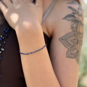 Pode incluir: Uma pulseira delicada com pequenas contas redondas azuis escuras. A pulseira &eacute; usada em um bra&ccedil;o, com uma tatuagem vis&iacute;vel na parte superior do bra&ccedil;o. A pulseira parece ser feita de pedras preciosas naturais.