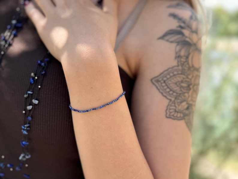 Pode incluir: Uma pulseira delicada com pequenas contas redondas azuis escuras. A pulseira &eacute; usada em um bra&ccedil;o, com uma tatuagem vis&iacute;vel na parte superior do bra&ccedil;o. A pulseira parece ser feita de pedras preciosas naturais.