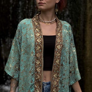 Könnte beinhalten: Ein offener Kimono in Blaugrün und Braun mit einem detaillierten Blumenmuster. Das Kleidungsstück hat einen breiten, dunkelbraunen Rand an den Vorderkanten, der mit einem sich wiederholenden goldenen und cremefarbenen Blumenmotiv verziert ist.
