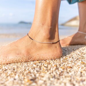 May include: A woman's foot with purple nail polish on her toes, wearing a black ankle bracelet with silver beads on a sandy beach.