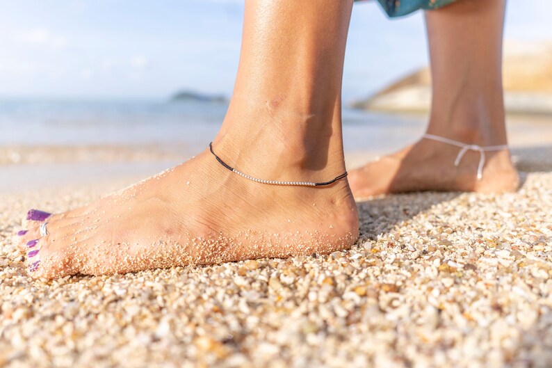 May include: A woman's foot with purple nail polish on her toes, wearing a black ankle bracelet with silver beads on a sandy beach.