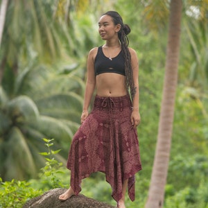 May include: A woman wearing a black sports bra and a long, flowing, burgundy skirt with a paisley pattern. She is standing in a tropical forest setting with palm trees in the background.
