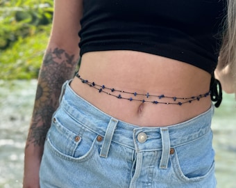 Pode incluir: Um cordão de contas de cintura de duas voltas com contas azul-escuro em um cordão preto. As contas são usadas em torno da cintura de uma pessoa que usa jeans azul.