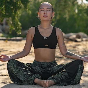 Könnte beinhalten: Eine Frau sitzt in meditativer Haltung auf einem Sandstrand, trägt einen schwarzen Sport-BH und eine schwarze und grüne gemusterte Hose. Sie hat die Augen geschlossen und ihre Hände sind in Mudra.