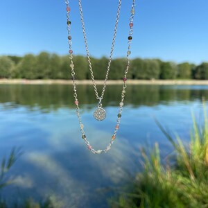 May include: A silver necklace with a delicate chain and a small, round pendant. The pendant is made of silver and has a geometric design. The necklace is layered with a second chain that has small, colorful beads.
