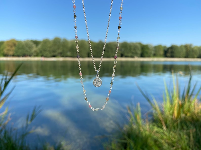 May include: A silver necklace with a delicate chain and a small, round pendant. The pendant is made of silver and has a geometric design. The necklace is layered with a second chain that has small, colorful beads.