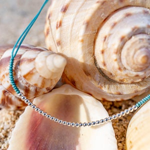 May include: A turquoise cord bracelet with silver beads, resting on seashells.