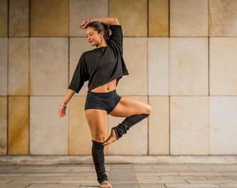 Pode incluir: Uma mulher com um top curto e calções pretos equilibra-se numa perna, com o braço levantado. Ela usa aquecedores de pernas pretos. O fundo é uma parede com painéis quadrados.