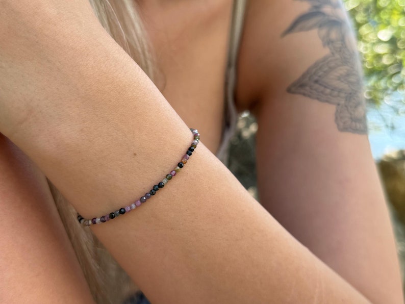 May include: A delicate bracelet made of small, multi-coloured beads. The beads, in shades of black, pink, and green, are strung together to create a slender band. The bracelet is worn on a wrist, with a blurred background of foliage.