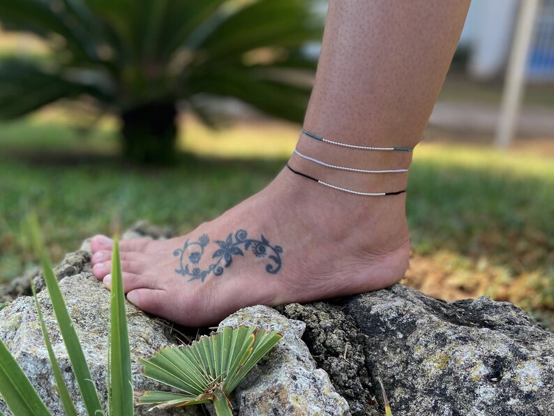 May include: A person's ankle with three anklets. The anklets are black, silver, and a light blue and green color. The person has a tattoo of a floral design on their ankle.