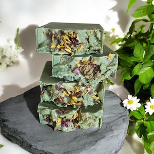 May include: A stack of four rectangular bars of sage green soap. Each bar features a textured surface embedded with dried yellow and purple flower petals. The soap bars are arranged vertically, showcasing a consistent color and botanical inclusions.