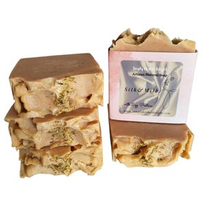 May include: Artisan natural soap bars in a stack and a half-wrapped bar. The bars are light brown with a textured surface and flecks of green. The label reads "Silk & Milk" with "Simply Herbal Delight" and "with Egg Protein".