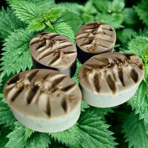 May include: Three oval-shaped bars of soap with a smooth, light brown surface featuring carved leaf-like indentations and a dusting of dried botanical particles on top, arranged against a background of vibrant green stinging nettle leaves.