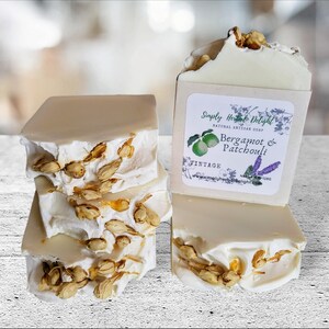 May include: Artisan soap bars with a light beige color and dried floral accents. One bar has a label that reads "Simply Herbal Delight" with "Bergamot & Patchouli" and a vintage design. The soap is stacked and arranged on a light-colored surface.