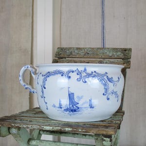 May include: A white ceramic chamber pot with blue nautical designs, including sailboats and decorative flourishes. The pot has a handle and sits on a weathered, light green wooden chair. The background features linen fabric with vertical blue stripes.