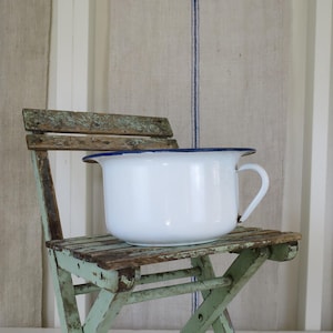 May include: A white enamel chamber pot with a blue rim and handle sits on a small, weathered, folding wooden chair. The chair has a distressed, faded green paint finish. The background features striped fabric panels.