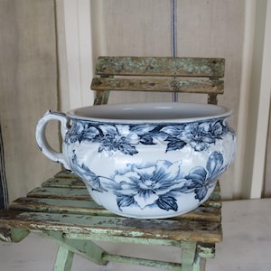 May include: A white and blue floral patterned chamber pot with a handle, resting on a small, weathered, green wooden chair. The floral design is in shades of blue. The background includes a striped curtain.