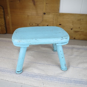 May include: A small, light blue wooden stool with a rectangular top and four rounded, slightly tapered legs. The paint finish shows signs of wear and distress, revealing the texture of the wood underneath on the edges and legs of the stool.