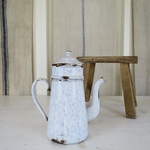 May include: A vintage white enamel coffee pot with a blue speckled pattern and a handle. The pot has a lid and a spout. A small wooden stool is next to the pot. The background features linen fabric.
