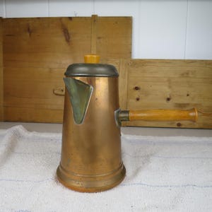 May include: A copper coffee pot with a cylindrical body, a side-mounted wooden handle, and a metal lid featuring a small yellow knob. The pot has a wide base and a short, angled spout. The metal surface shows signs of age and natural patina.