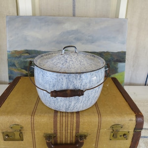 May include: A vintage enamelware pot with a lid, featuring a blue and white speckled pattern. It has a metal handle and wooden grip. The pot is sitting on a vintage suitcase with brown leather trim.