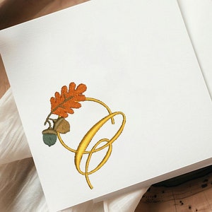 Peut inclure: Une carte blanche avec un motif de bague dorée et une feuille orange et verte avec des glands. La carte est sur une surface en bois avec un tissu blanc.