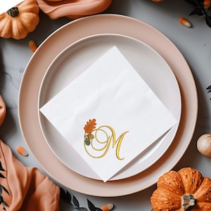May include: A festive table setting with a white napkin featuring a gold monogram "M" and autumn leaf design. The napkin rests on a pink plate, surrounded by orange pumpkins, peach-colored fabric, and decorative elements, creating a fall theme.