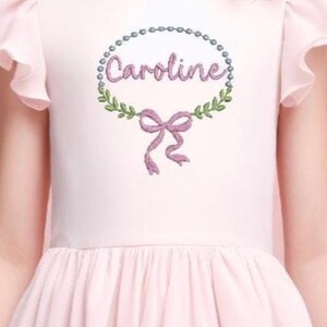 Könnte beinhalten: Hellrosa Kleid mit rundem Ausschnitt und kurzen, gerüschten Ärmeln. Das Kleid hat eine Stickerei auf dem Oberteil mit dem Namen "Caroline", umgeben von einem Kranz und einer Schleife.