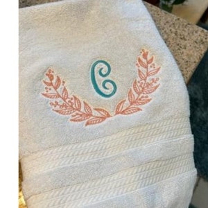 Puede incluir: Una toalla blanca con una letra C bordada en verde turquesa con una corona de hojas de color melocotón. La toalla tiene el texto "Pine Tree Embroidery" debajo del monograma.