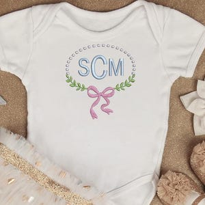 Puede incluir: Un body blanco para bebé con monograma y lazo rosa, combinado con una falda tutú con lunares dorados y zapatos a juego. Estrellas decorativas y un collar de perlas rodean el conjunto, creando un tema caprichoso.