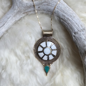 May include: A gold chain necklace with a round pendant featuring a white and gold sun design. The pendant has a turquoise teardrop hanging from the bottom.