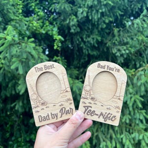 May include: Two wooden golf-themed picture frames. One frame reads "The Best Dad by Par" and the other says "Dad You're Tee-rific". Each frame has a circular opening for a photo and golf-related illustrations.